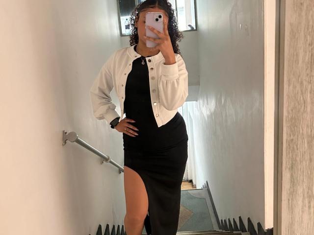 SofiaPassionne, with curly hair, wearing a white jacket and black dress with a thigh slit, taking a mirror selfie, showing her hand on her hip, and a raised phone.