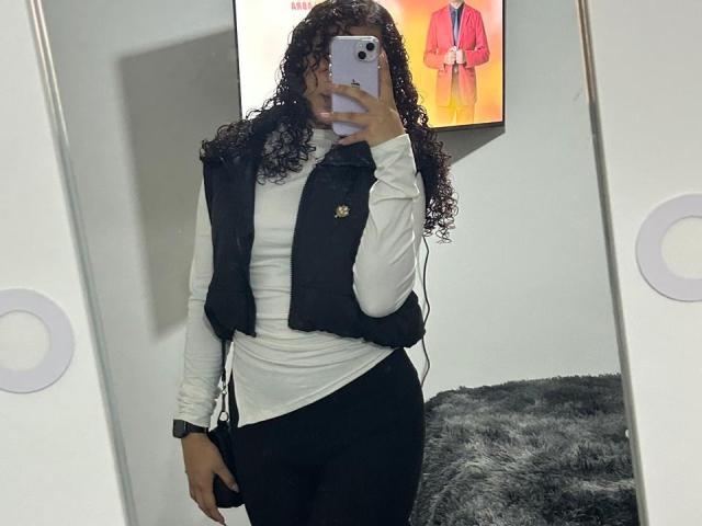 SofiaPassionne with curly dark hair, wearing a white long-sleeve shirt, black vest, and black pants, taking a mirror selfie, holding a purple phone, showing her butt.