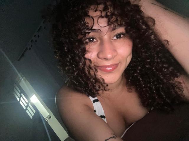 Sofia, passionnée, avec ses cheveux bruns bouclés, souriante, montrant ses lèvres roses brillantes et sa poitrine dans un débardeur rayé, levant un bras, dans une pièce faiblement éclairée..