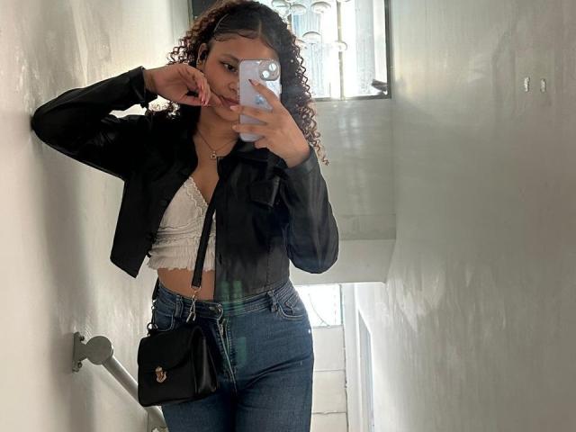SofiaPassionne with curly brown hair, black jacket, white lace bra, and high-waisted blue jeans, holding a phone, wearing a black crossbody bag, standing in a hallway.