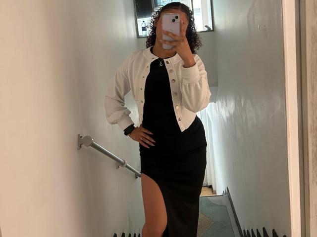 Sofia a les cheveux bouclés, porte une veste blanche, une robe noire fendue au niveau des cuisses, et tient un téléphone dans sa main. Elle prend un selfie devant un miroir dans un couloir..