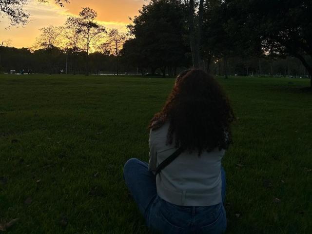 Sofia, passionnée, avec des cheveux bruns bouclés, une chemise blanche et un jean bleu, assise sur l'herbe, dos à la caméra, regardant le coucher de soleil orange, des arbres sombres en arrière-plan, une sangle en travers du torse..