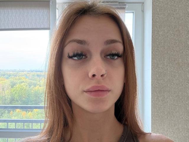MarisolRothery has light brown hair, blue eyes, long eyelashes, a nose ring, and pink lips. She's wearing a gray top, looking serious with a slight pout.