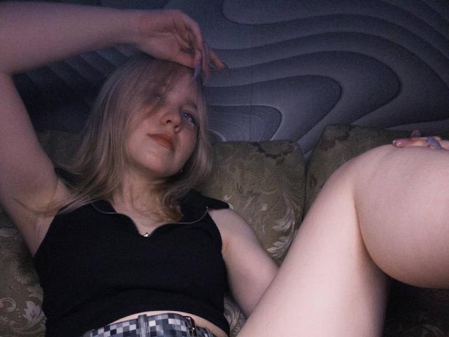 TiffaniMacandog, con i capelli biondi, un top corto nero e una gonna a quadri, sdraiata su un divano, un braccio sopra la testa, le gambe divaricate, che mostra cosce chiare, con un'espressione rilassata e un sottile rossetto rosso..