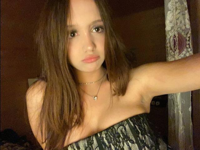 NuPastiva with long brown hair, wearing a strapless black-and-white patterned top, has small tits, a subtle necklace, and a serious expression, standing in a dimly lit room.