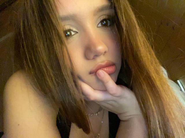 NuPastiva with long brown hair, biting her finger, looking at the camera with slightly parted lips, green eyes, and a necklace, in a dimly lit room.