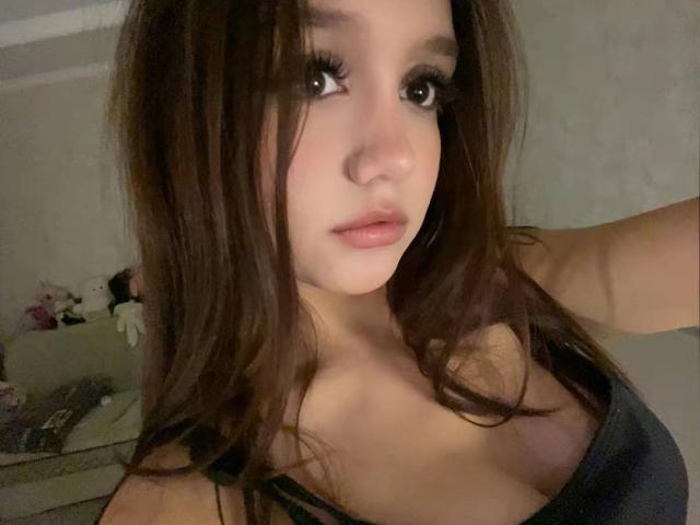NuPastiva has long brown hair, big green eyes, and pink lips. She's wearing a dark tank top that shows her small tits. Her right arm is raised.
