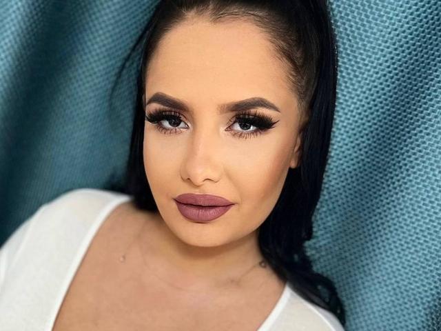 SabelaLyn has thick black eyelashes, dark brown eyebrows, full pinkish-brown lips, and a light tan complexion. She's wearing a white top with long black hair.