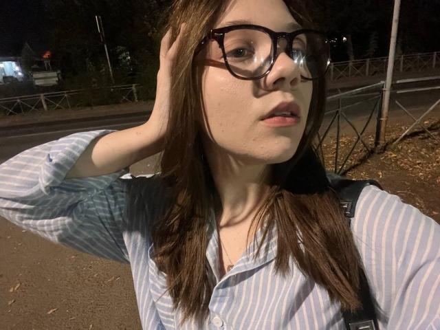 AureliaEmswiler with glasses, striped shirt, brown hair, hand in hair, looking at camera, slight pout, backpack, night, street, tree shadows, soft light, casual selfie.