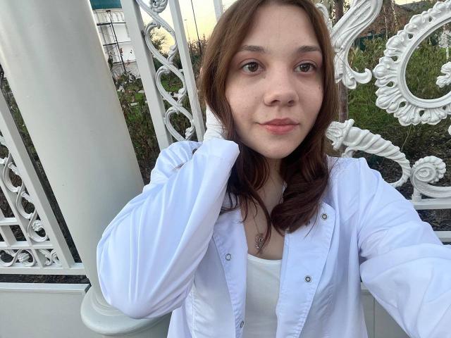 AureliaEmswiler with brown hair, white shirt, and white jacket, holding her hair, looking at the camera, showing her small, cute tits, and a subtle smile.