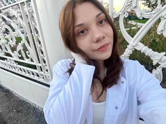 AureliaEmswiler with brown hair, wearing a white shirt, has a tattoo under her collarbone, standing by a white ornate fence, looking at the camera with a slight smile.
