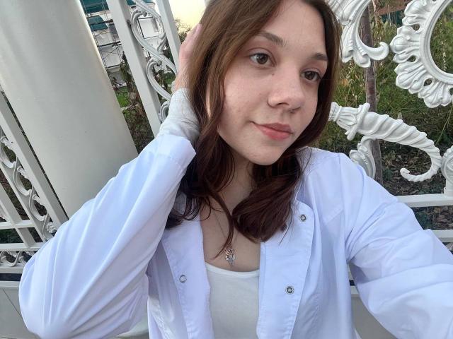 AureliaEmswiler with brown hair, wearing a white shirt and white tank top, has her hand in her hair, showing a slight smile, and a necklace with a small pendant.