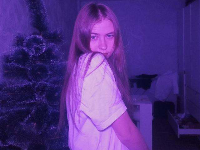 MargretWarriner with long, straight, red-brown hair, wearing a white shirt, looking over her shoulder with a serious expression, standing next to a purple-lit Christmas tree.