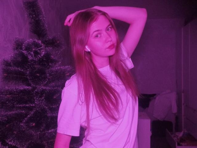 MargretWarriner with long red hair, white shirt, arm raised, looking at camera, standing next to a decorated Christmas tree, bathed in pink light.