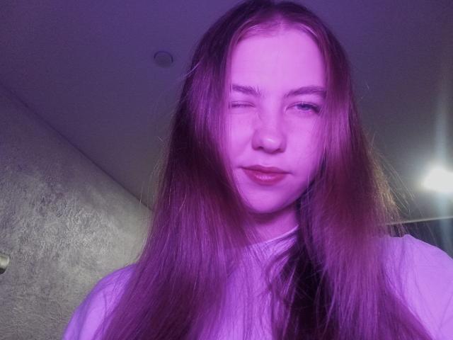 MargretWarriner with long purple-tinted hair, winking, and a slight smile, showing her pouty ass-kissing lips. She's wearing a white shirt, with a subtle light source behind.