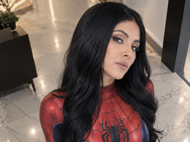 MaddieDemie with long black hair, wearing a red Spider-Man suit, looking at the camera with a slight smile, showing her makeup and blue eyes, standing in a shiny room.