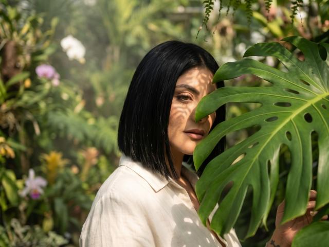 MaddieDemie with black bob, holding a large green monstera leaf, wearing a white shirt, sunlight casting shadows on her face, surrounded by lush greenery.