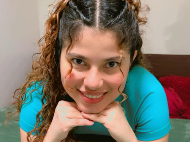 EmilyCM with curly brown hair, blue shirt, smiling, hands under chin, looking at camera, cute face, medium tits, light skin, brown eyes, red lipstick, relaxed, indoor setting.