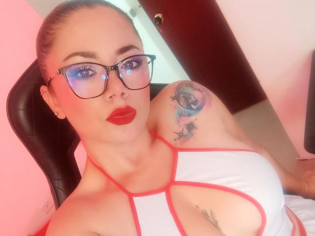SamanthaVenus with glasses, red lipstick, white and red top, large boobs, tattoo on right shoulder, pink walls, black chair, blonde hair in ponytail, looking at camera.