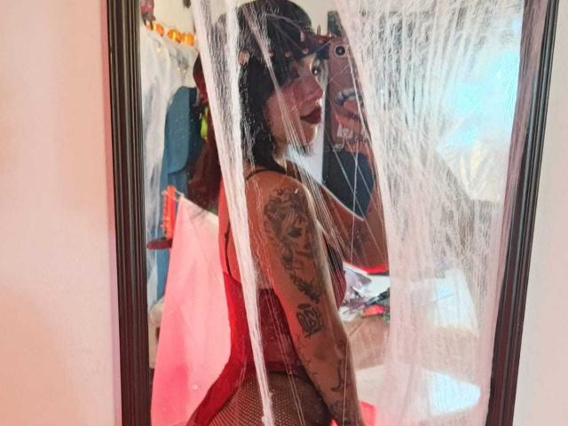 DominicTheOne with dark hair and red lipstick, showing a tattooed arm, wearing fishnet stockings, reflected in a mirror with white webbing, standing in front of a pink bag.