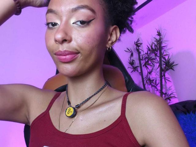 KiaraKamau hat ein hübsches Gesicht mit pinkem Lippenstift, schwarzen Eyeliner-Akzenten und einem roten Tanktop. Sie trägt eine schwarze Halskette mit einem gelben Smiley und kleinen goldenen Ohrringen..