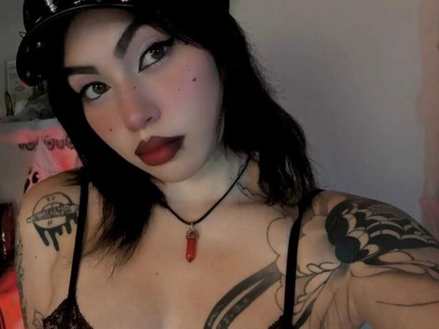 DominicTheOne with black hair, dark eyeliner, red lipstick, black tank top, tattoos on arms and chest, red crystal necklace, black hat, looking at camera, pale skin, full breasts.