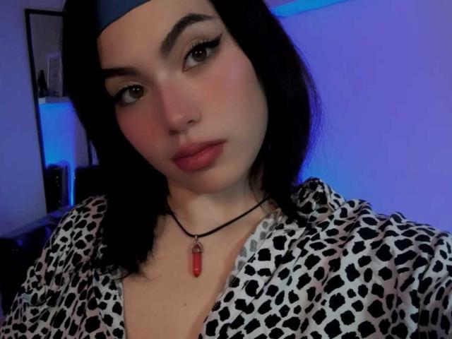 DominicTheOne with black hair, blue headband, eyeliner, and red lipstick, wearing a leopard-print shirt, necklace with red pendant, and a neutral expression. Blue lighting in background.