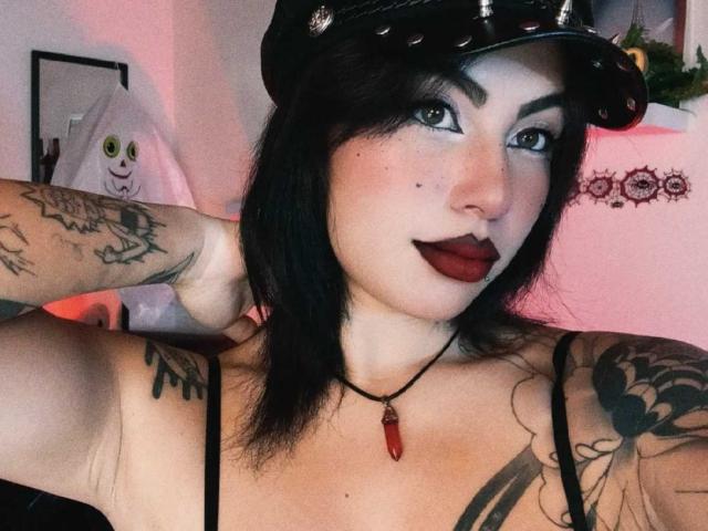 DominicTheOne with black hair, dark lipstick, tattoos, and a black cap, wearing a red crystal necklace, showing tits and arms. Pinkish room lighting. Smirking at the camera.