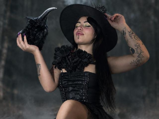 DominicTheOne with black feathers in hand, wearing a black corset and hat, has tattoos and green eyeshadow, pouting with nose ring, holding a black crow.