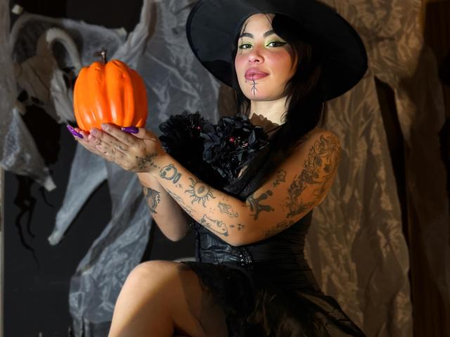 DominicTheOne with tattoos, holding a bright orange pumpkin, wearing a black witch hat and dress, has colorful green eye makeup, and a black floral necklace, in a dark setting.