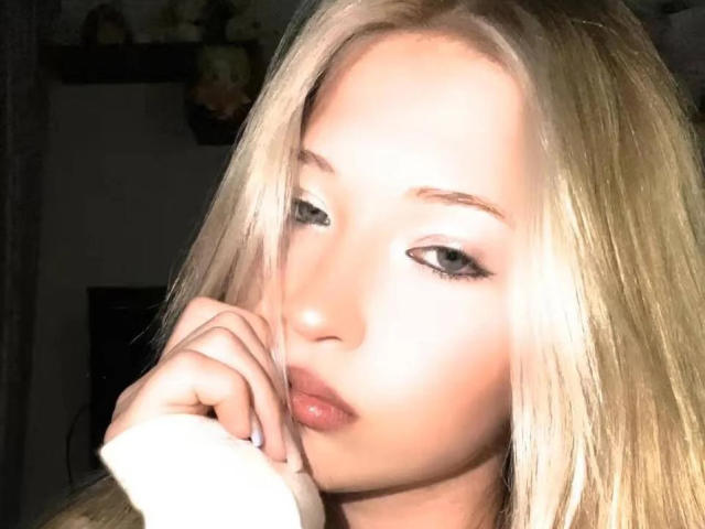 LadyLuu with blonde hair, fair skin, and full lips, pouting with a hand near her mouth, has sultry eyes and a slight smile, looking directly at the camera.