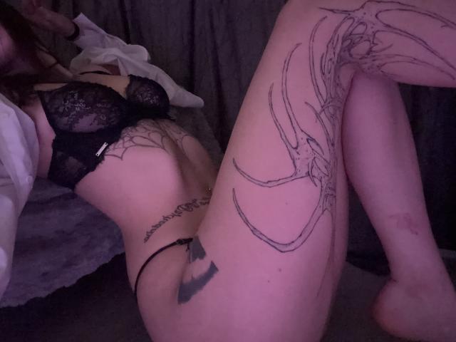Simonne Brustkern, con un sujetador y tanga de encaje negro, acostada en la cama, con las piernas levantadas, mostrando un gran tatuaje de telaraña en el muslo y un tatuaje en el trasero, con una iluminación púrpura tenue..