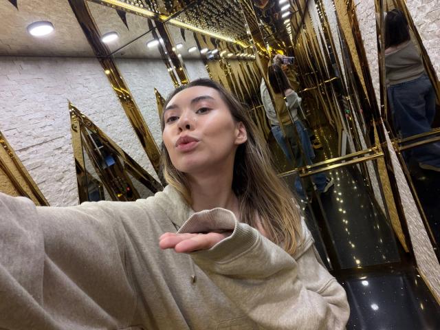 GwenOnline with pouty lips, light brown hair, and beige sweater, hand out, wearing a gold mirror wall and white brick background. Her ass is not visible.