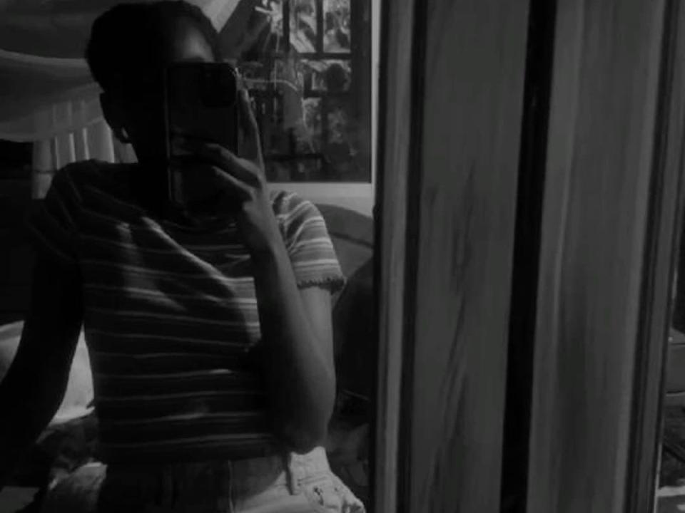 Black and white selfie of CocoGustavo69 with dark skin, in a striped shirt, holding a phone, covering their face, reflected in a mirror, showing their upper body and arm.