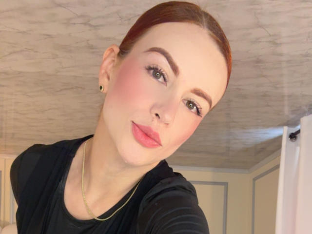 Leahred a les cheveux roux, les yeux verts, un blush rose et du rouge à lèvres rose. Elle porte une chemise noire et un collier en or, avec un léger sourire et un anneau de nez mignon..
