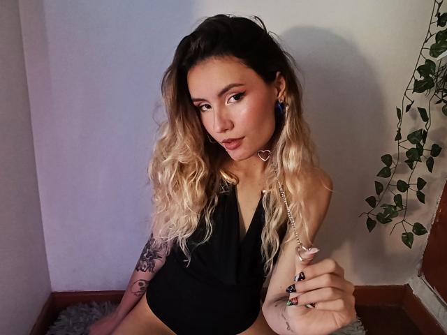 Alanis Blanc, com cabelo loiro e ondulado, top preto de alça, colar com pingente de coração, tatuagens, segurando um brinquedo sexual rosa, olhando para a câmera, sentada encostada em uma parede, com uma planta ao fundo..