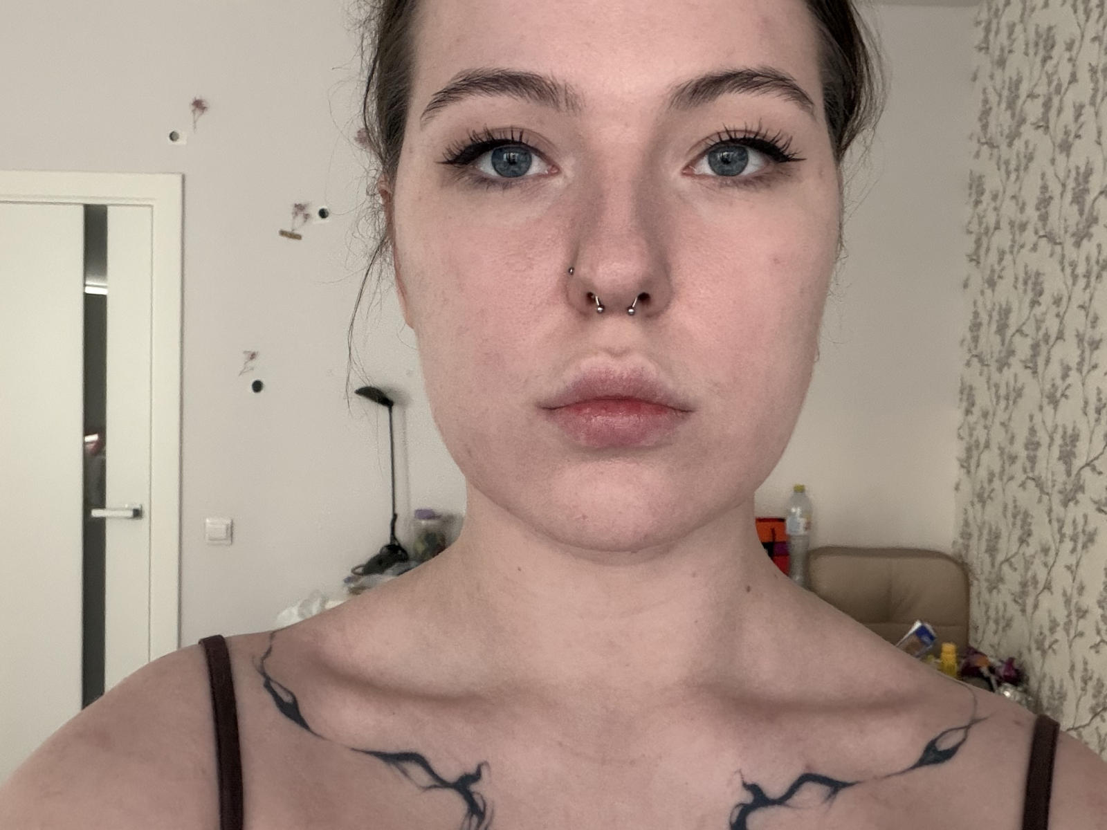 Deneen Angelone heeft bleke huid, blauwe ogen, zwarte eyeliner, een neusring en zwarte tatoeages op haar sleutelbeenderen. Ze kijkt recht in de camera met een neutrale uitdrukking..