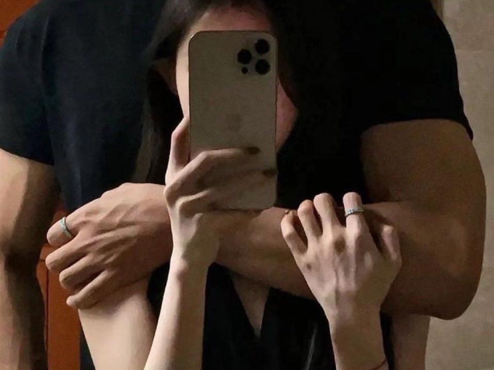 SandraFunny with long black hair, holding a phone in front of her face, arms around a muscular guy in a black shirt, his arms around her, in a mirror selfie.