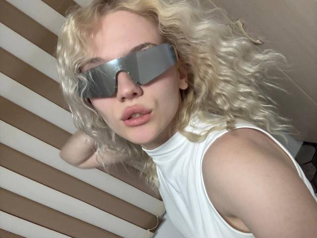 YungReher with curly blonde hair, wearing a white sleeveless top and reflective silver visor, pouting with slightly parted lips, in a casual selfie. Boobs not visible.