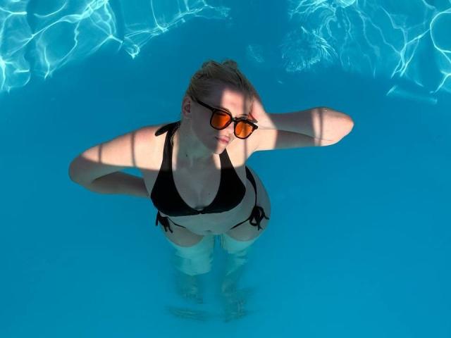 YungReher in a black bikini, wearing orange sunglasses, with hands behind head, standing in clear blue water, looking up. Her tits are visible, slightly submerged.