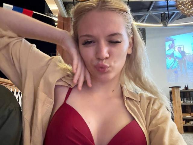 Blonde YungReher with pouty lips, wearing a red bra and open beige shirt, has her hand on her cheek, showing off her tits. Blonde hair, light skin.