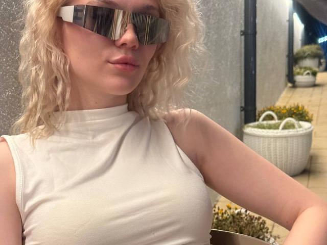 YungReher with curly blonde hair, wearing reflective sunglasses, and a white sleeveless top, showing small tits, in a slightly blurred selfie.