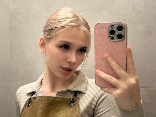 YungReher with blonde hair in a ponytail, wearing a beige shirt and brown apron, holding a pink iPhone, taking a mirror selfie, showing her pouty lips and subtle makeup.