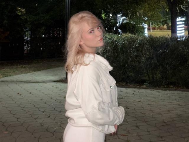YungReher with blonde hair, white fluffy jacket, and white pants, standing sideways, looking back, hands in front, on a brick-paved path at night.