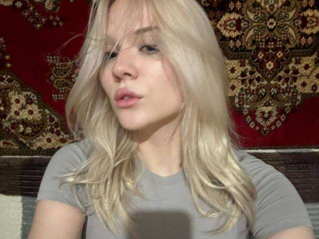 YungReher with blonde hair, pouty lips, and light skin, wearing a grey tee, has a cute face and small tits. She's looking at a phone in a selfie.
