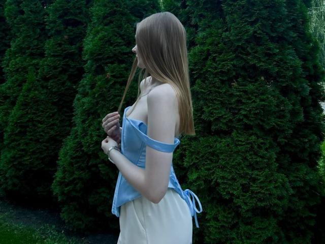 Solange Gulsvig ha lunghi capelli rossi, indossa una maglietta blu con una spalla scoperta che mette in mostra il suo seno piccolo, e una gonna bianca, ed è in piedi di fronte a cespugli verdi alti, tenendo in mano un bastoncino sottile..