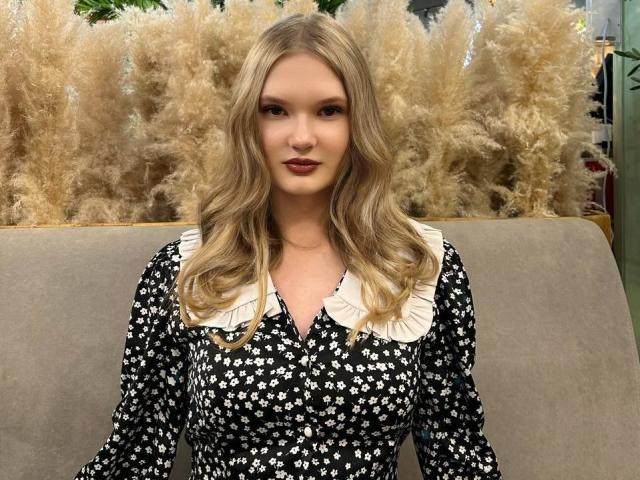 Solange Gulsvig a les cheveux blonds et ondulés, elle porte une robe noire avec des motifs floraux blancs et un col blanc frangé, et elle se tient devant de hautes herbes de pampa..
