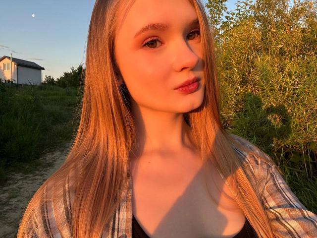 Solange Gulsvig, avec de longs cheveux blonds, une peau claire et des lèvres pulpeuses, portant une chemise à carreaux, baignée de lumière du soleil, regardant la caméra, debout sur un sentier herbeux, montrant ses seins..