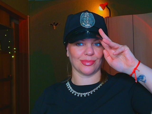 VeronikaGoldy in a police cap, saluting with right hand, has blue eyes, pink lipstick, black shirt, pearl necklace, red bracelet, and a tattoo on her left wrist.