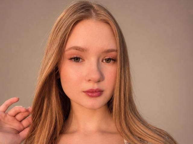 SolangeGulsvig has long blonde hair, green eyes, and pink lips. She's looking straight at the camera with a slight, confident smile, holding her hair on the left.